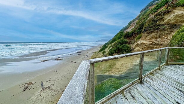 Une terrasse en bois avec des rampes de protection en verre, donnant sur une plage bordée d’une falaise et d’un océan.