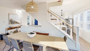 Eine moderne Küche mit einem Edelstahl-Kühlschrank, ein Essbereich mit einem Holztisch und Korbstühlen sowie eine Treppe mit Glastrennwand, die in ein Obergeschoss führt.