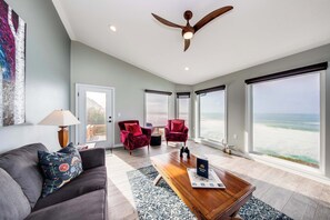 Un salon spacieux doté d’une grande fenêtre donnant sur la plage, d’un ventilateur de plafond et d’une table basse en bois sur laquelle est posé un magazine.