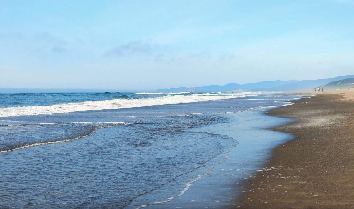 A beach with waves lapping onto the shore, a clear sky, and distant mountains.
