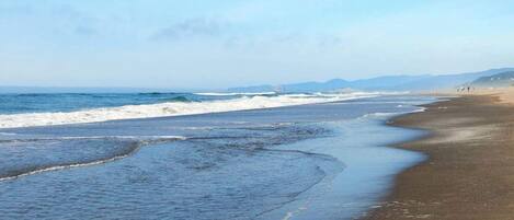 A beach with waves lapping onto the shore, a clear sky, and distant mountains.