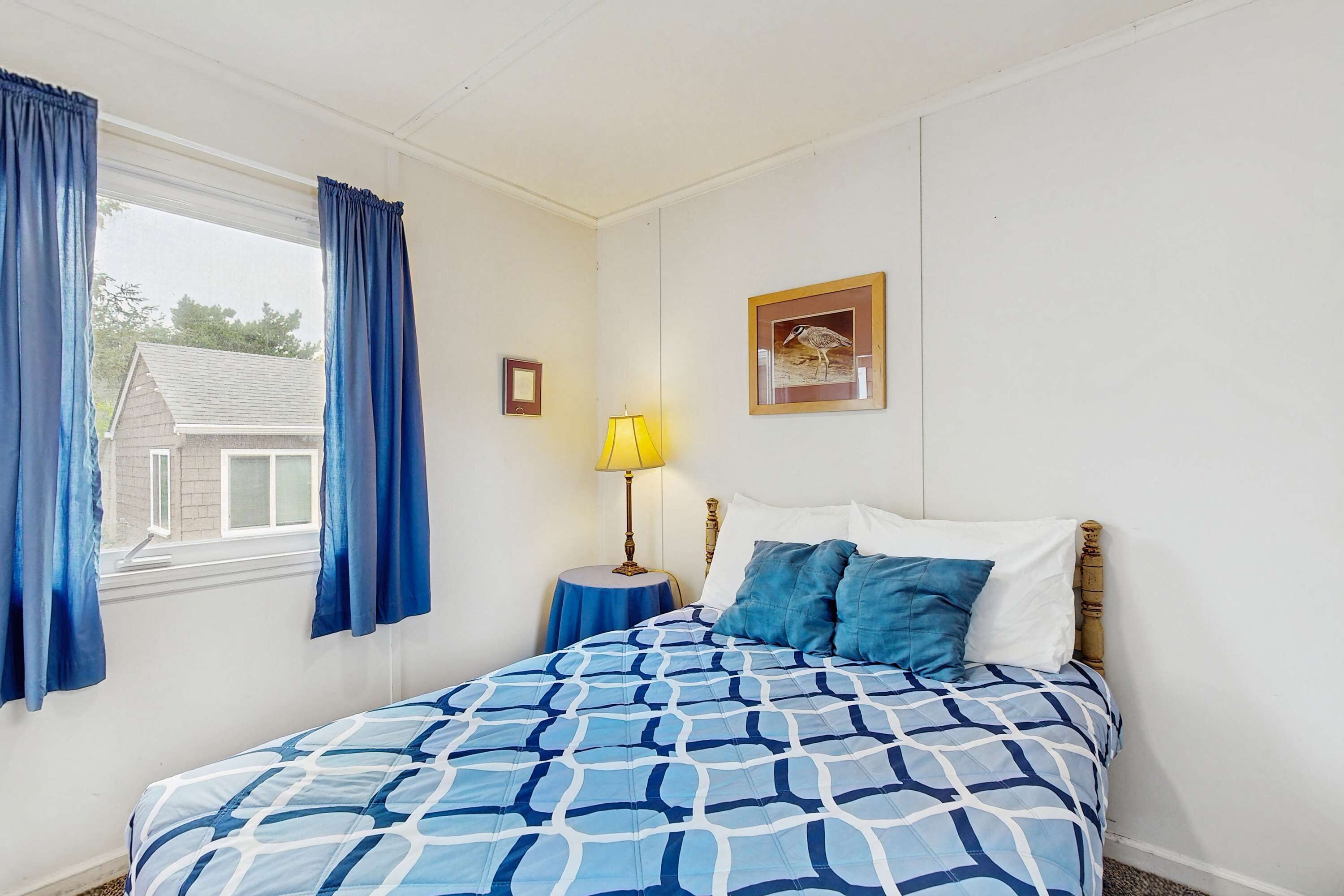 Un dormitorio con cama, ventana con cortinas, mesita de noche con lámpara y un cuadro enmarcado en la pared.