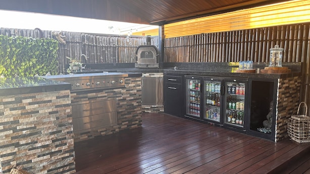 Ein Außenküchenbereich mit einer Steinarbeitsplatte, einem Grill und einem mit Getränken gefüllten Kühlschrank.
