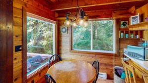 A wooden cabin interior with a dining table, chairs, a kitchen area, and large windows offering a forest view. - Dog friendly cabin with ocean view fast WiFi fireplace and washer dryer (Otis)