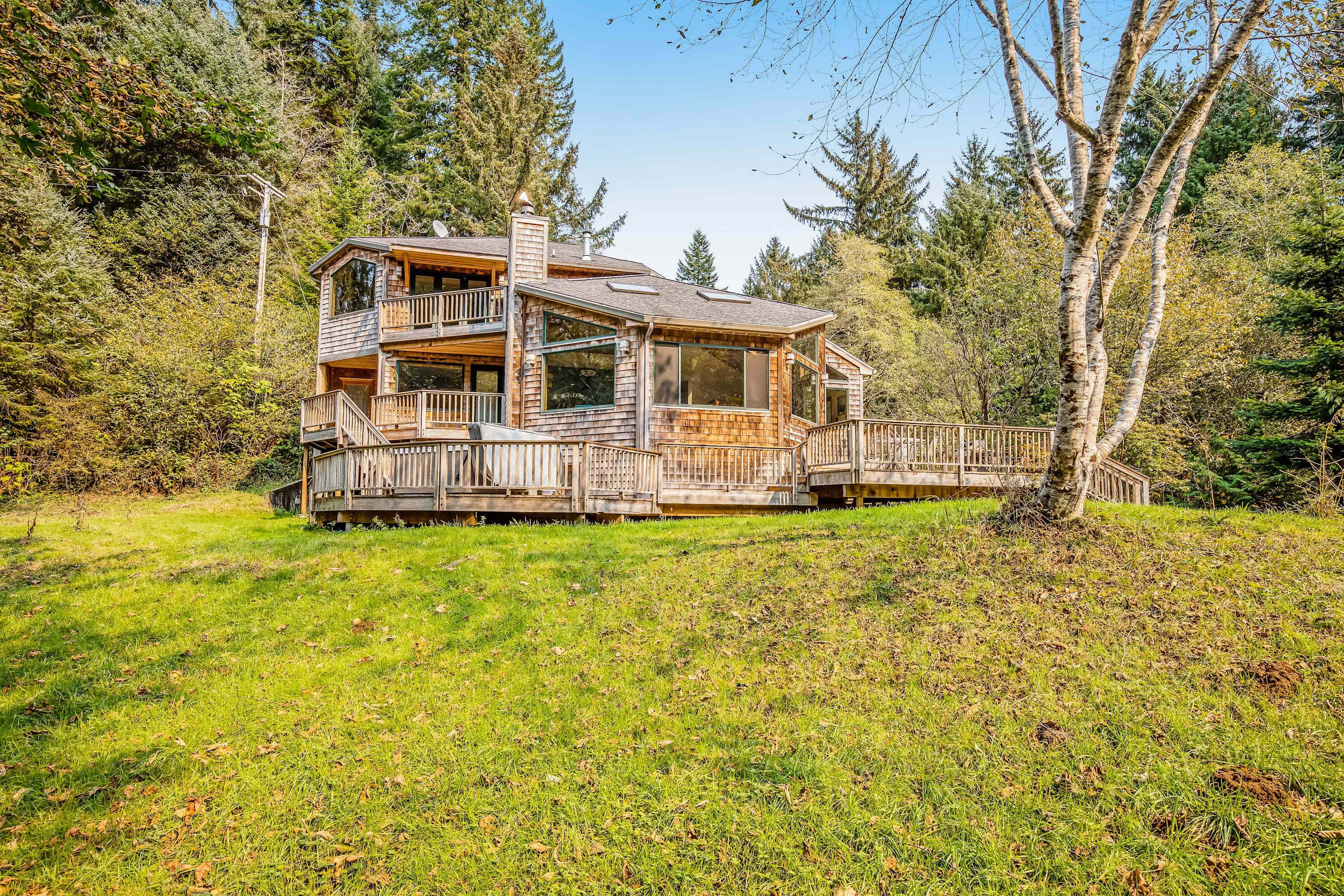 A two-story wooden cabin with a deck, surrounded by trees and a grassy area.