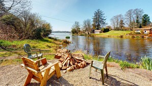 Un cadre idyllique au bord du lac, avec des bancs en bois et un foyer, le tout entouré d’arbres et sous un ciel dégagé.