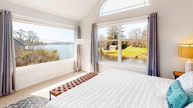 Une chambre avec un grand lit, deux fenêtres avec des rideaux, une vue sur un lac et un espace extérieur, ainsi qu’une lampe de chevet.