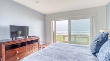 Une chambre avec un lit, un téléviseur posé sur une commode en bois, et une grande porte-fenêtre coulissante donnant sur un balcon avec vue sur l’océan.