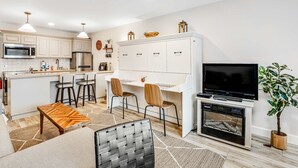 A modern kitchen with white cabinets, a microwave, and a stainless steel refrigerator. There is a dining area with a white table and chairs, and a TV mounted on the wall.
