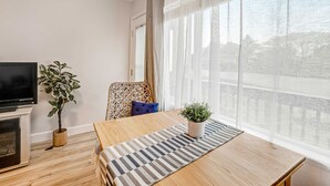 A modern living room with a wooden dining table, a potted plant, a television, and a large window with sheer curtains.