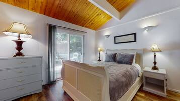 A bedroom with a wooden ceiling, a bed with a headboard, two bedside tables with lamps, a nightstand, a dresser, and a window with a view of greenery.