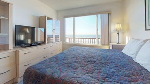 Una camera d'hotel con un letto grande, una televisione su una cassettiera, un comodino con una lampada e vista sull'oceano da ampie finestre.