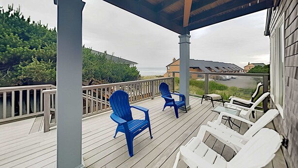 Eine überdachte Terrasse mit blauen und weißen Adirondack-Stühlen, einem Tisch und Blick auf Strand und Meer.