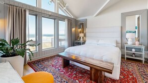 Habitación de hotel con una cama grande, un escritorio, una silla, un tapete rojo estampado, una planta en maceta y vistas a la playa a través de amplias ventanas.