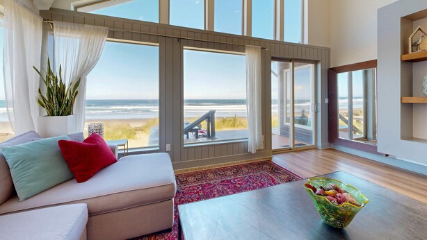Un salón espacioso con un amplio ventanal con vista a la playa, un sofá acogedor con cojines, una mesa de centro de madera y una planta decorativa.