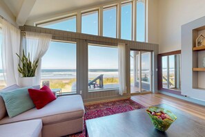 A spacious living room with a large window overlooking a beach, a cozy sofa with cushions, a wooden coffee table, and a decorative plant. - New oceanfront home with hot tub views A C and beach access 2 dogs OK (Neskowin)