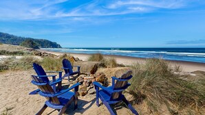 Escena de playa con cuatro sillas Adirondack azules dispuestas alrededor de una fogata, con vista a una playa arenosa y el océano.