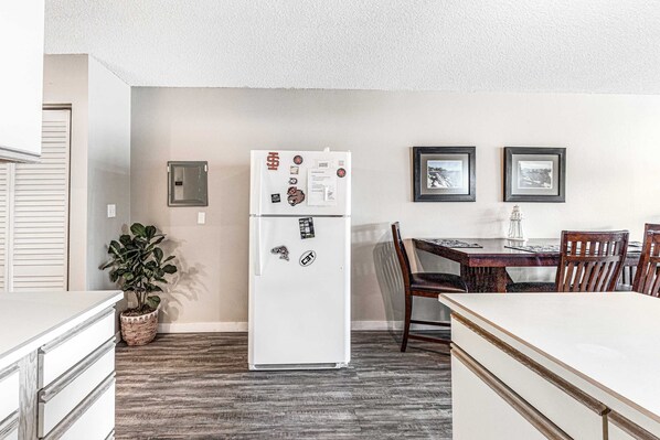 A compact kitchen with a refrigerator, dining table, chairs, and a potted plant. - Oceanfront 2nd floor condo with river ocean views balcony (Neskowin)