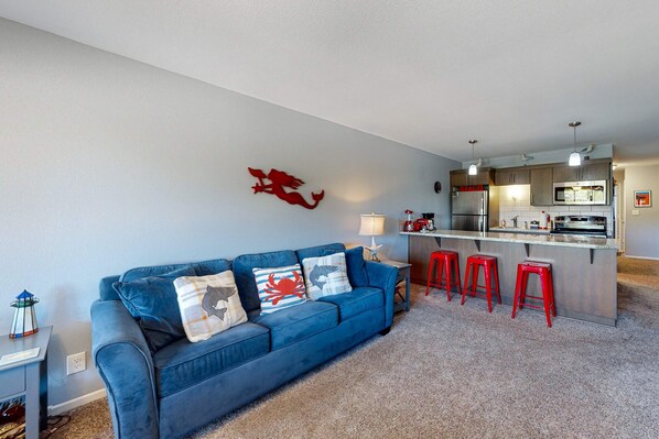 A living room with a blue sofa, a kitchen area, and a red bar stool. - Third floor beachfront condo with a balcony full kitchen and ocean views (Neskowin)