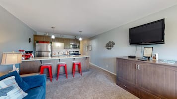 A compact living space with a kitchenette, a blue sofa, red bar stools, and a flat-screen TV mounted on the wall.