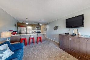 A compact living space with a kitchenette, a blue sofa, red bar stools, and a flat-screen TV mounted on the wall. - Third floor beachfront condo with a balcony full kitchen and ocean views (Neskowin)