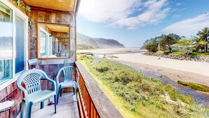 Un balcon avec deux chaises, une balustrade en bois et une vue sur une plage, avec une colline boisée en arrière-plan.
