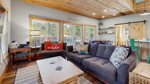 A cozy living room with a wooden ceiling, a grey sofa, a wooden coffee table, and a kitchen area in the background. - Newly constructed home with firepit and large deck walk to the beach and golf (Neskowin)