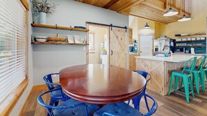 A dining area with a wooden table and blue chairs, a kitchen with a white refrigerator and a sink, and a wooden ceiling with exposed beams. - Newly constructed home with firepit and large deck walk to the beach and golf (Neskowin)