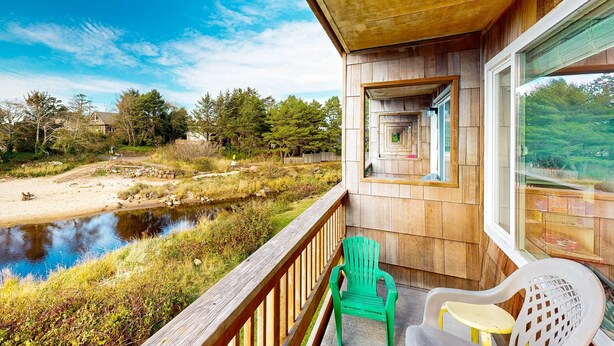 A wooden deck with a view of a river and forest, a small outdoor seating area with chairs, and a window with a view of the landscape. - Creekside condo with beach access balcony full kitchen and ocean views (Neskowin)
