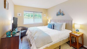Un dormitorio con cama, televisor sobre un mueble, ventana y una obra de arte colgada en la pared.