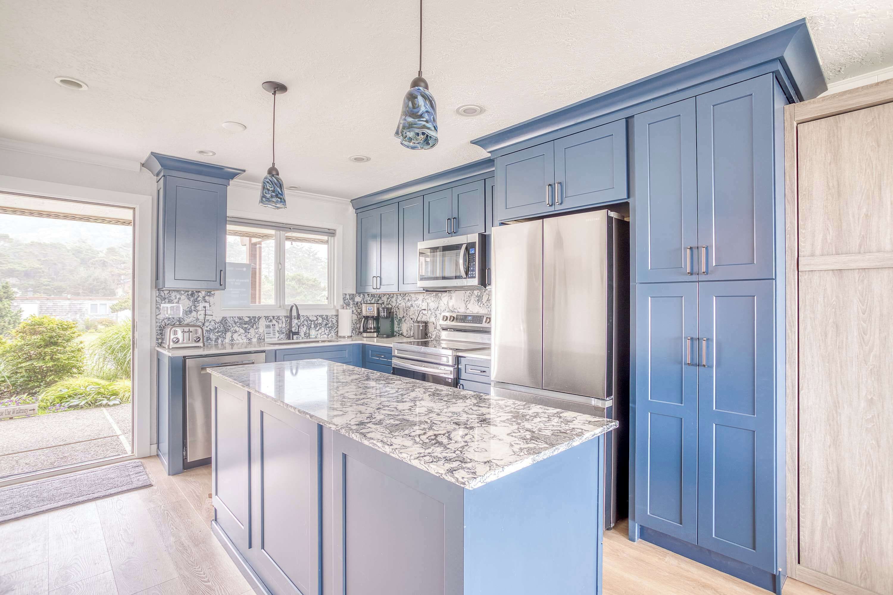 A modern kitchen with blue cabinets, a marble island, stainless steel appliances, and pendant lighting.