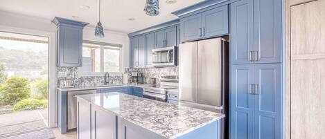 A modern kitchen with blue cabinets, a marble island, stainless steel appliances, and pendant lighting.