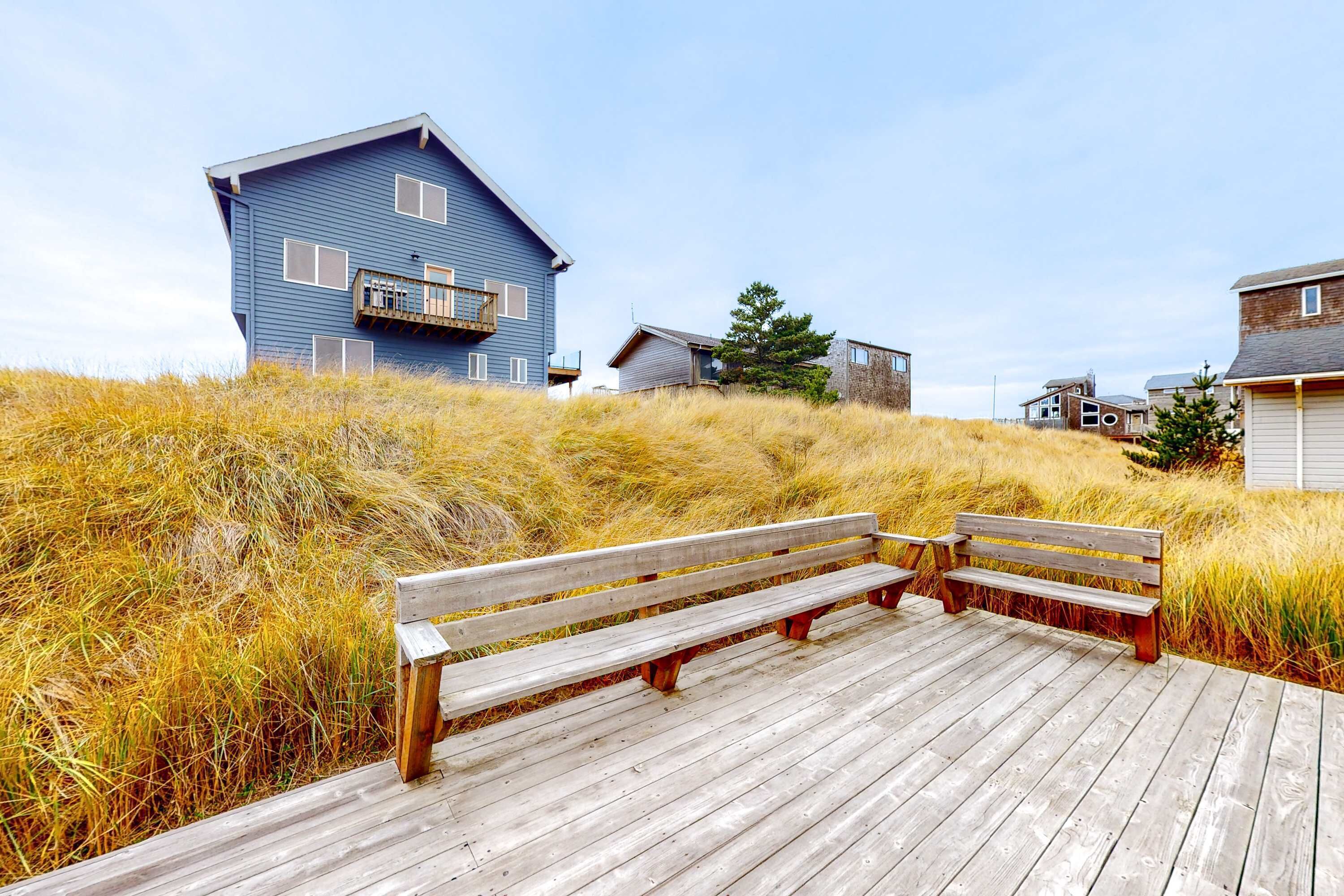 A wooden deck with benches, a blue house, and surrounding grassy terrain.