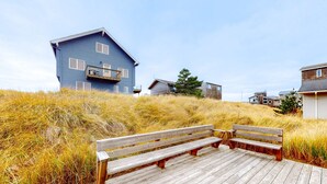 Una terraza de madera con bancos, una casa azul y terreno cubierto de césped.