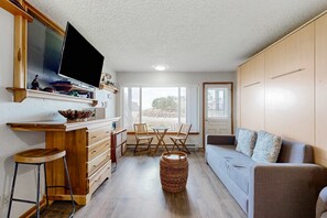A compact living space with a kitchenette, a TV mounted on a wooden cabinet, a dining area with a table and chairs, and a sofa with cushions. - Beachfront dog friendly studio with amazing Proposal Rock view steps to the sand (Neskowin)