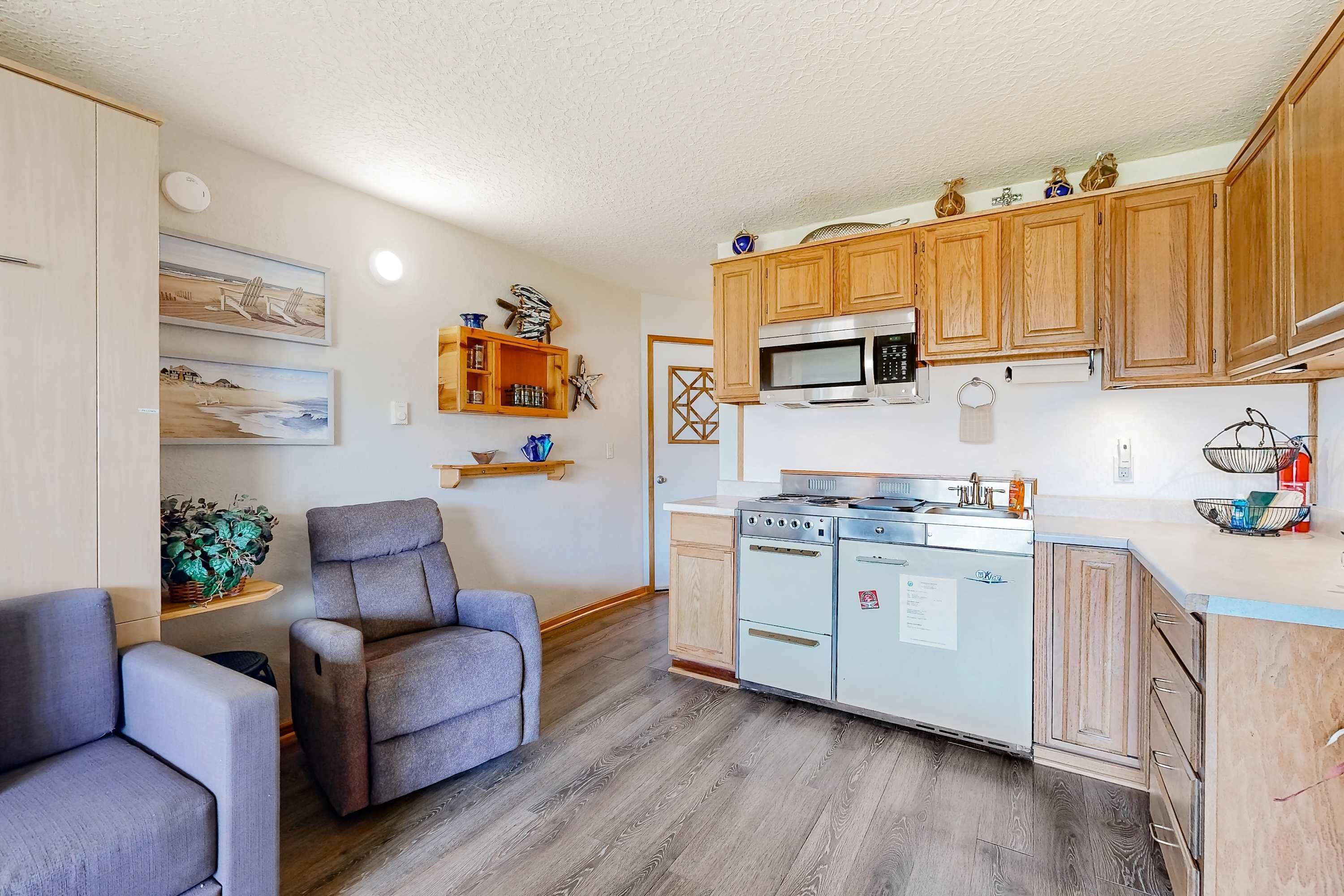 A compact kitchen with wooden cabinets, a microwave, and a stove. There is a gray chair and a small shelf with decorative items.