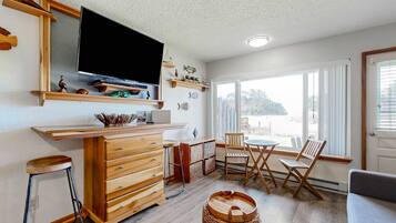 A cozy room with a wooden dresser, a TV mounted on the wall, a dining area with chairs and a table, and a large window offering a view of the outdoors.