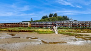A multi-story building with a wooden facade and balconies, situated near a natural landscape with a clear sky. - Oceanfront 2nd floor condo with ocean views balcony and seasonal beach access (Neskowin)