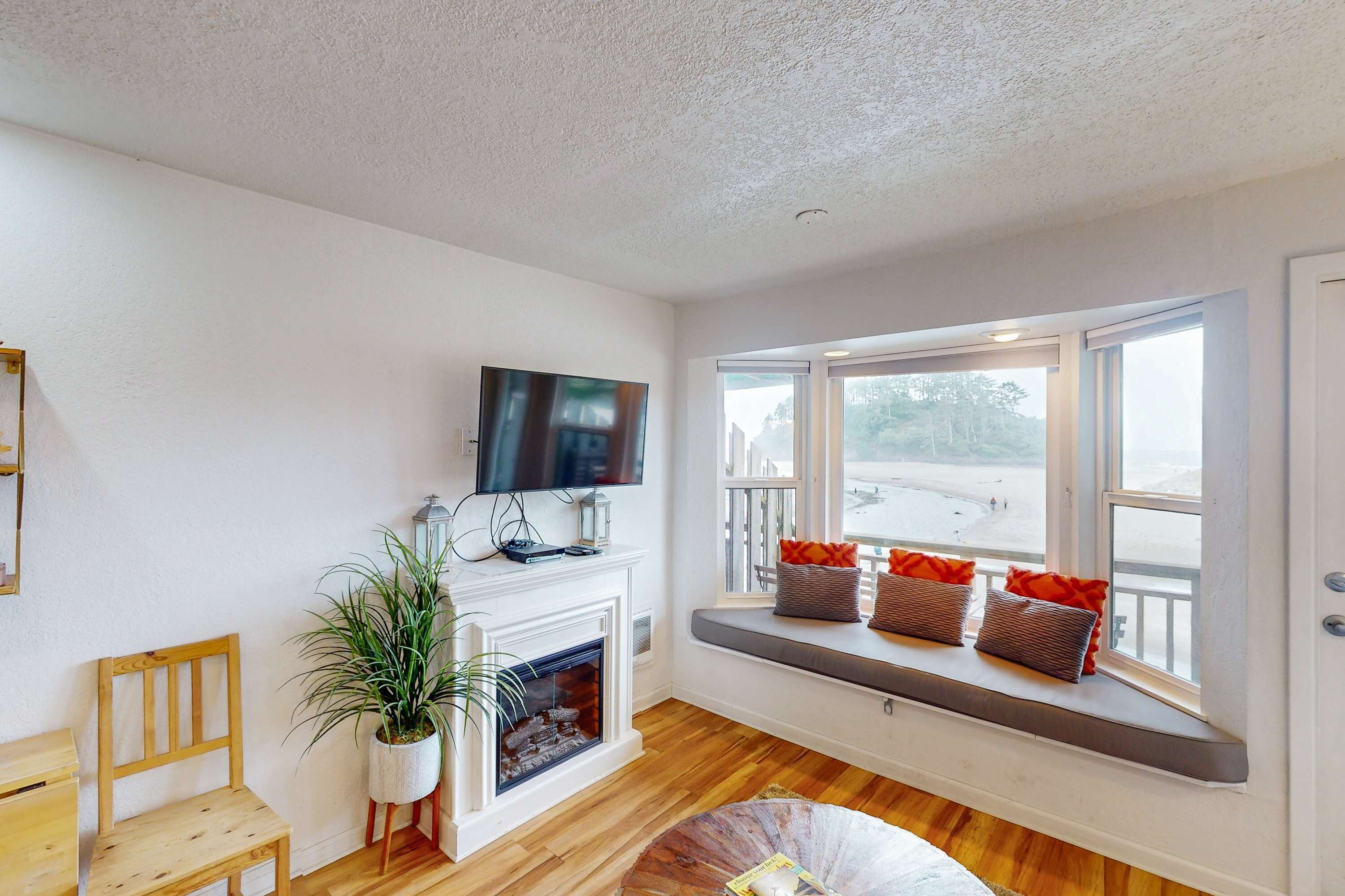 A living room with a fireplace, a television mounted above it, a bench with cushions by a large window, and a wooden chair.