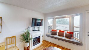 A living room with a fireplace, a television mounted above it, a bench with cushions by a large window, and a wooden chair. - Oceanfront 2nd floor condo with ocean views balcony and seasonal beach access (Neskowin)