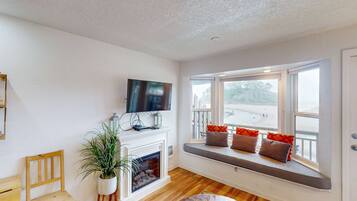 A living room with a fireplace, a television mounted above it, a bench with cushions by a large window, and a wooden chair.