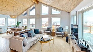 Un salon spacieux avec un plafond haut, de grandes fenêtres, un plafond en bois, un coin repas avec une table et des chaises, et un canapé avec des coussins.