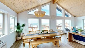 Une pièce spacieuse avec un plafond en bois, de grandes fenêtres, une table à manger avec des bancs, et un coin salon comprenant un canapé et une table d’appoint.