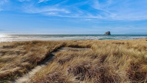 Ein Küstenpfad mit trockenem Gras, der zum Meer führt, ein Felsvorsprung in der Ferne und ein klarer, blauer Himmel.