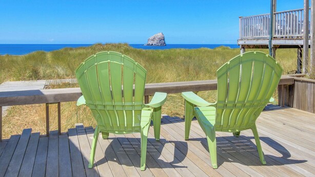 Zwei grüne Adirondack-Stühle auf einer Holzterrasse mit Blick auf einen Strand und einer Felsformation in der Ferne.