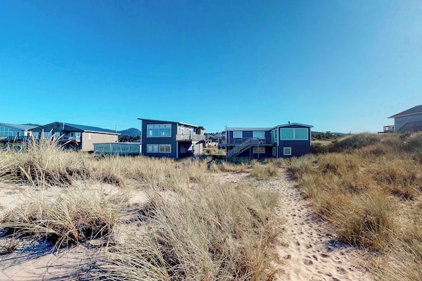 A beachfront property with modern houses, a sandy path, and surrounding dunes. - Beautiful oceanfront home with a gorgeous view (Pacific City)
