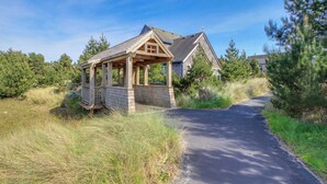 Ein hölzerner Gazebo mit geneigtem Dach, ein gepflasterter Weg, der zu einem Haus mit Satteldach führt, umgeben von Bäumen und Gras.