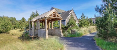 A wooden gazebo with a sloped roof, a paved pathway leading to a house with a gabled roof, surrounded by trees and grass.