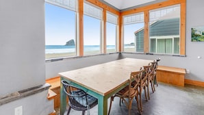 Ein Essbereich mit großem Tisch und Stühlen, großen Fenstern mit Holzrahmen sowie Blick auf Strand und Meer.