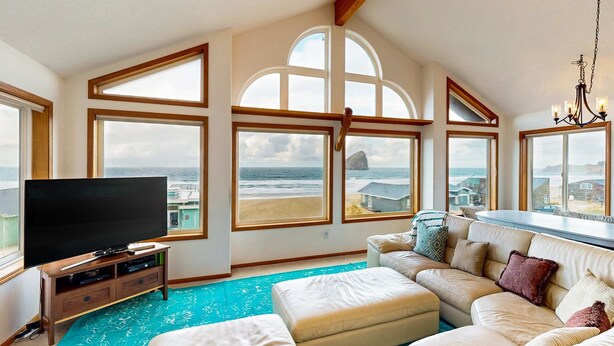 A spacious living room with a large flat-screen TV, a beige sectional sofa, and a view of the beach through large windows. - Gorgeous waterfront home with ocean views a furnished deck and beach access (Pacific City)